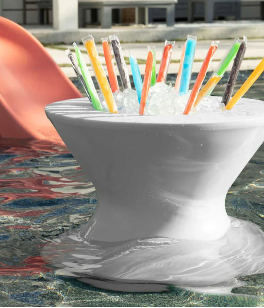 A white floating cooler filled with ice and colorful freeze pops sits in a pool near a red slide, with buildings in the background.