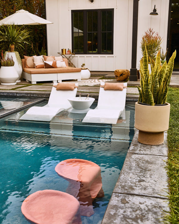 Outdoor poolside setup with two white loungers partially submerged in the pool, each with a rose-gold pillow. A round side table between them holds a drink. Nearby, an umbrella shades a seating area, and a potted plant is on the stone border.