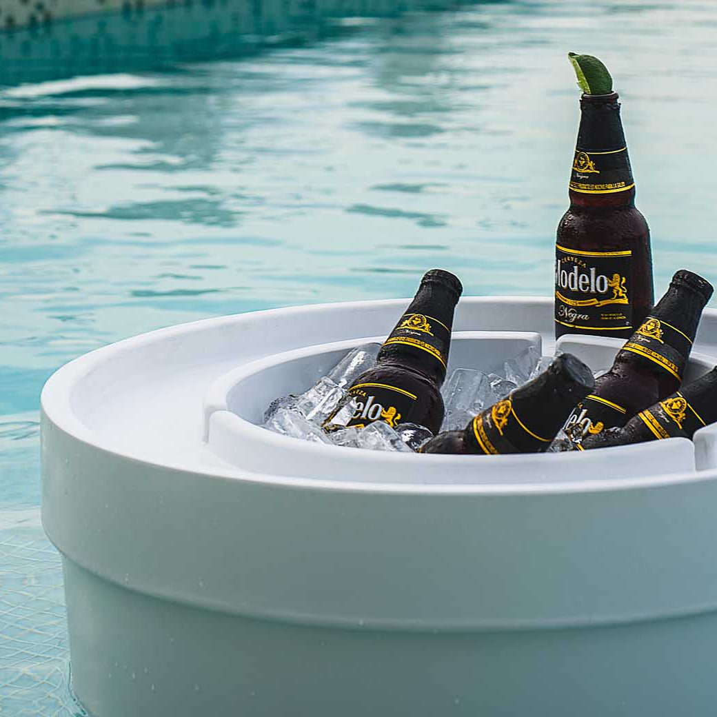 Five bottles of Modelo beer chill in a Signature Ice Basin by Ledge Lounger, filled with ice and floating on a clear blue swimming pool. One bottle stands upright with a lime wedge on the rim, while sunlight reflects off the water, creating a relaxing summer ambiance.