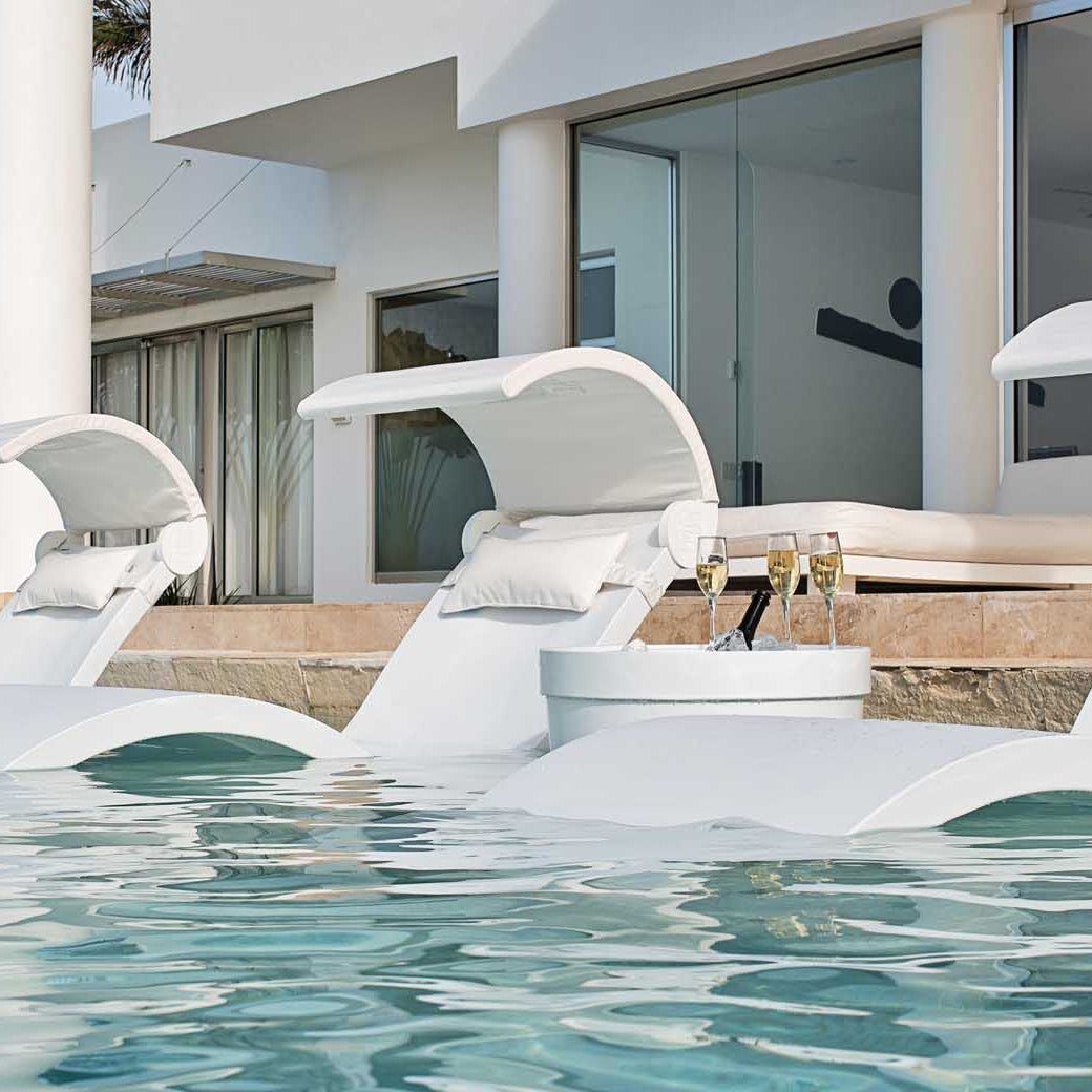 Three white lounge chairs are submerged in a modern pool outside a contemporary home with large glass doors. A small table, featuring the Signature Ice Basin by Ledge Lounger with a bottle and glasses, enhances the serene setting. Palm trees and a chaise lounge complete the scene.
