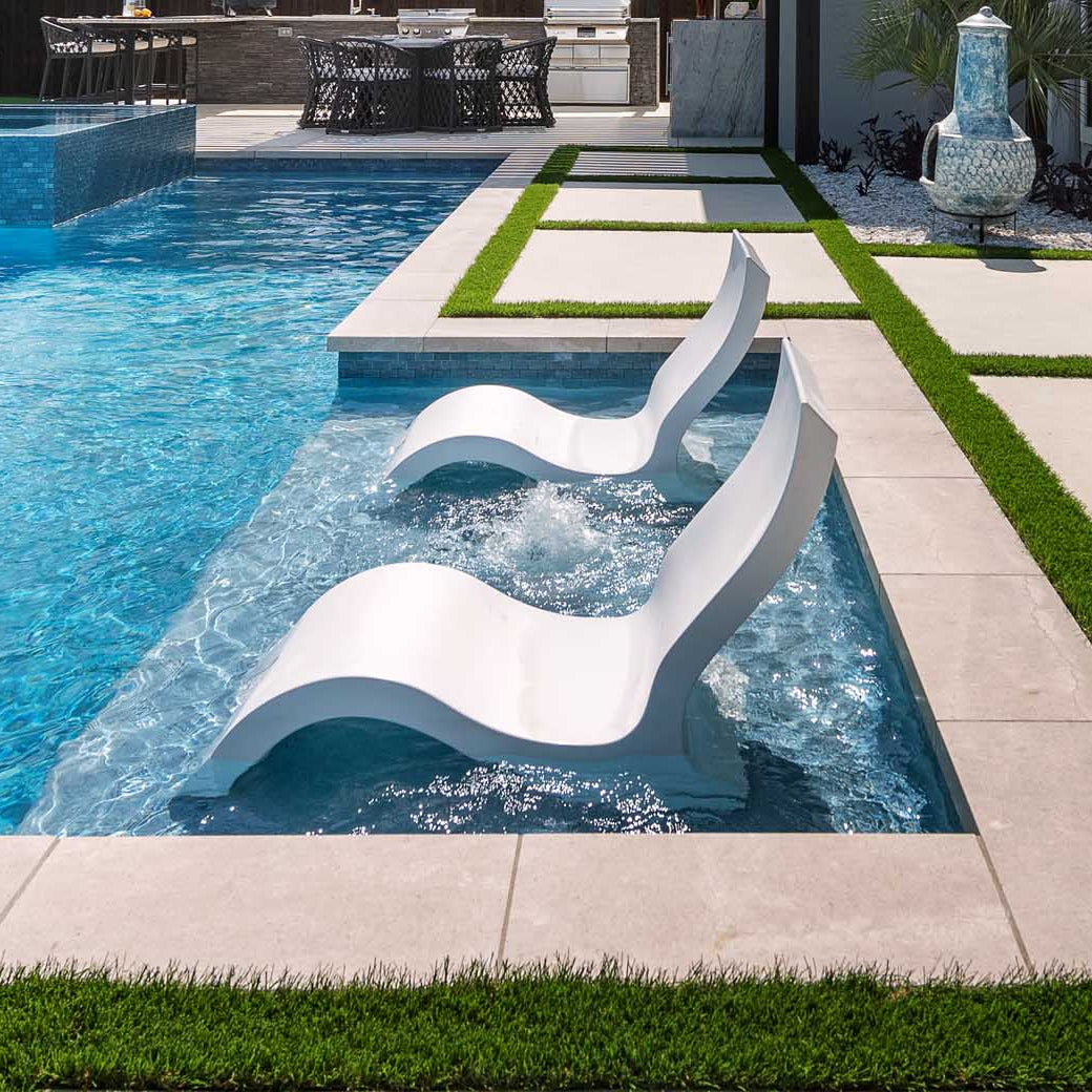 Two Ledge Lounger Signature Lowback Chairs are partially submerged in a modern pool, forming a serene outdoor oasis. A stone patio with artificial grass borders the area, while sunlight dances on the clear blue water beside a large decorative vase.