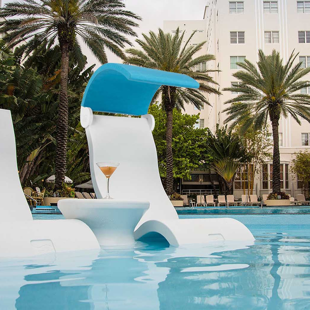 Two Signature Chairs by Ledge Lounger rest on a tanning ledge, partially submerged, with a small table holding a cocktail. Tall palm trees and a white building form the backdrop, creating an outdoor oasis with a tropical and relaxed atmosphere.