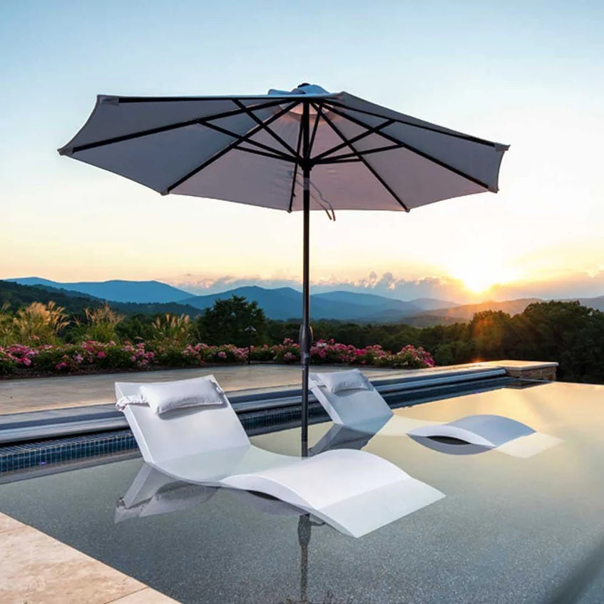 Two white lounge chairs sit in shallow pool water beneath a large umbrella, overlooking a scenic landscape of mountains and greenery at sunset.