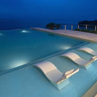 At dusk, the infinity pool features a tanning ledge with three submerged Signature Chaise Deep lounge chairs by Ledge Lounger, offering a tranquil ocean view. The pool edge merges seamlessly with the horizon as soft lighting enhances the serene ambiance.
