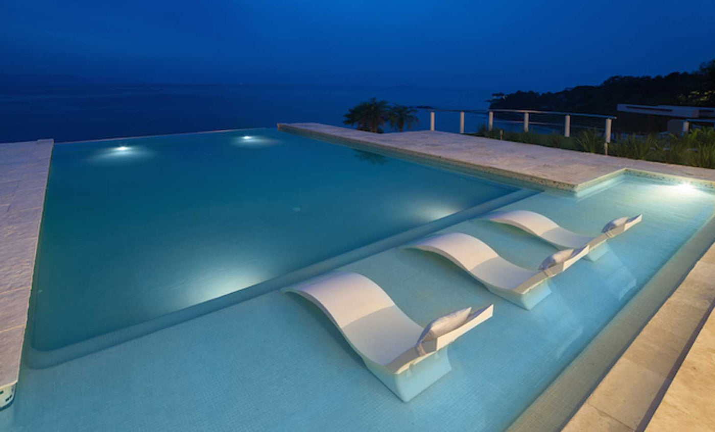 At dusk, the infinity pool features a tanning ledge with three submerged Signature Chaise Deep lounge chairs by Ledge Lounger, offering a tranquil ocean view. The pool edge merges seamlessly with the horizon as soft lighting enhances the serene ambiance.