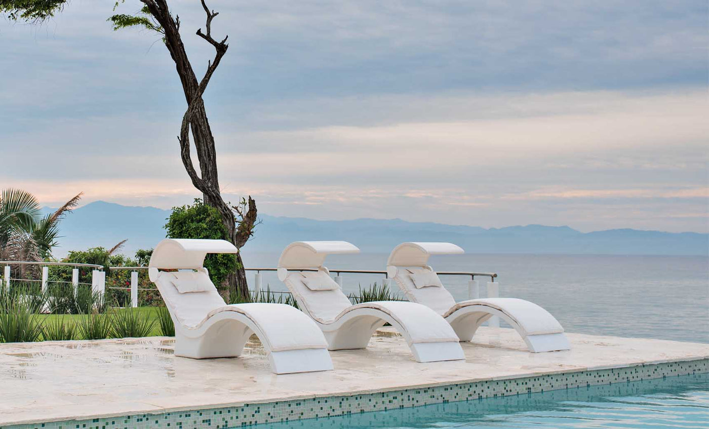 Three Signature Chaise Deep lounges by Ledge Lounger sit poolside, overlooking a tranquil ocean under a cloudy sky. A solitary tree is to the left with greenery on the horizon, creating a serene setting ideal for relaxing on the tanning ledge.