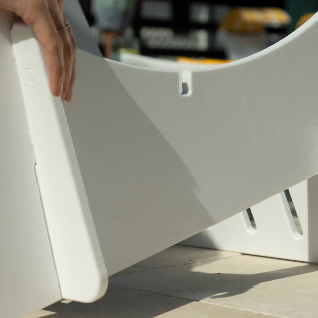 A person is assembling a white cardboard structure, fitting slotted pieces together like a Signature Lowback Chair Riser by Ledge Lounger. The scene is brightly lit with various blurred objects in the background.