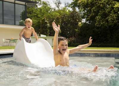 Two children playing in a pool with a slide, surrounded by greenery.