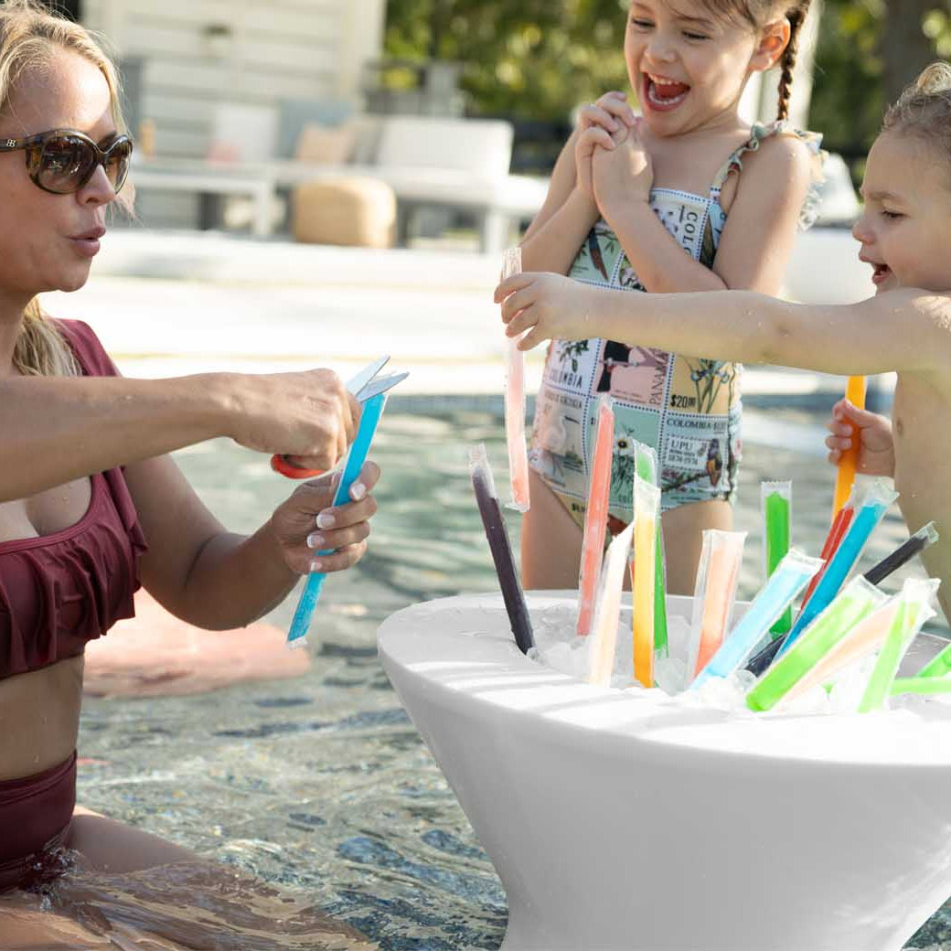 A woman in a maroon swimsuit stands in a pool with two children, selecting colorful ice pops from a Ledge Lounger Signature Tall Ice Bin Side Table. The girl wears a patterned swimsuit and the boy, swim trunks. Ledge Lounger Umbrellas provide shade over the lively patio area in the background.