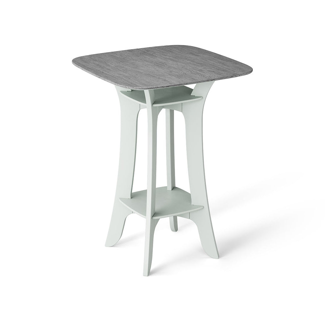 The Haven Hightop Table by Ledge Lounger is a modern square table with a grey top and light green, ergonomically curved legs. It features a small shelf below the tabletop, offering unique style and functionality as part of our patio collection.