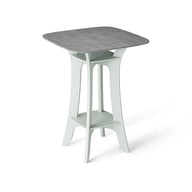 The Haven Hightop Table by Ledge Lounger is a modern square table with a grey top and light green, ergonomically curved legs. It features a small shelf below the tabletop, offering unique style and functionality as part of our patio collection.