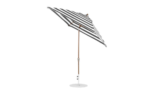 A Ledge Lounger black-and-white striped umbrella, the Essential Fiberglass Umbrella - 7.5 Octagon Crank Auto Tilt, stands upright with a wooden pole on a white base. Constructed from marine-grade fabrics, this wind-resistant umbrella tilts slightly to reveal its striking stripe pattern.