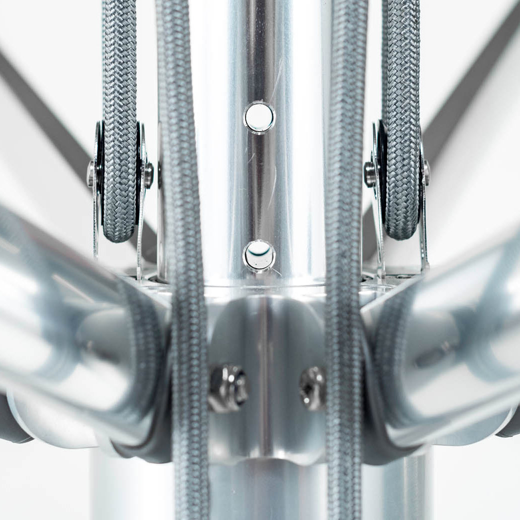 Close-up of a metallic structure with shiny cylindrical tubes converging at a central point. Gray straps attach to the tubes, enhancing the designs wind resistance. The blurred background highlights its symmetry and industrial appeal, reminiscent of a Ledge Lounger Premier Aluminum Umbrella.