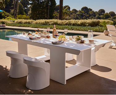 A modern white outdoor dining table set with white chairs, styled with food and wine glasses on a patio beside a swimming pool and landscaped garden.