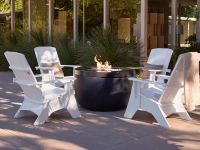 Four white outdoor Adirondack-style chairs arranged around a round black fire pit table with visible flames, set on a patio in front of a modern glass building with plants.