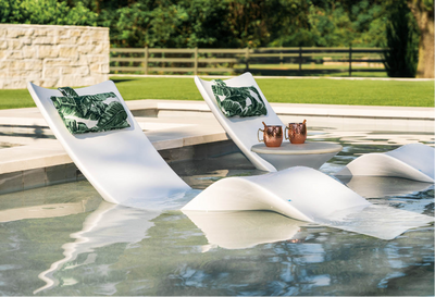 Modern white outdoor lounge chairs with green tropical leaf patterned pillows, set in a swimming pool next to a small side table with two copper mugs, with a grassy lawn and trees in the background.