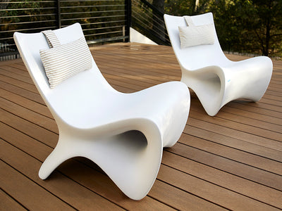 Two modern white outdoor lounge chairs with curved designs on a wooden deck. Each chair has a striped cushion at the top. The background features a railing and greenery in the distance.