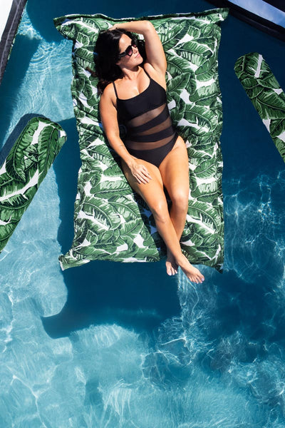 A woman in a black swimsuit lounges on a large, green tropical leaf-patterned float in a clear blue pool. She is wearing sunglasses and has one arm resting above her head.