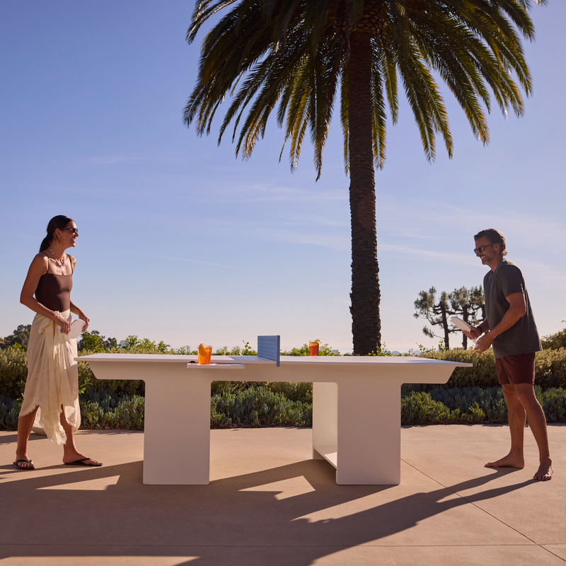 Two people play table tennis outdoors on a modern white table, with palm trees and a clear sky in the background. Orange drinks sit on the table, and both players are casually dressed.