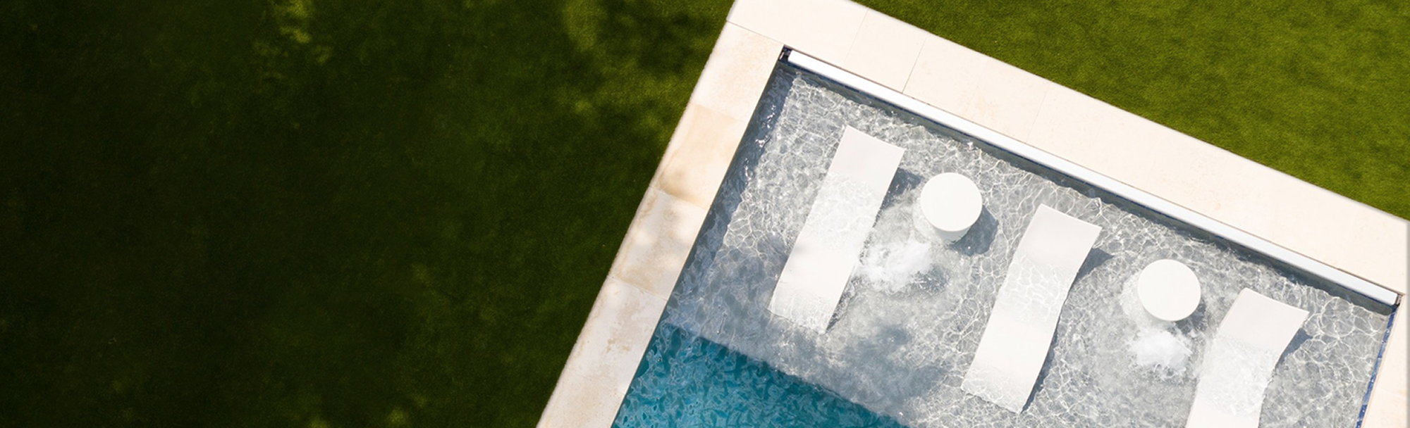 Aerial view of three white lounge chairs and round side tables partially submerged in the shallow end of a swimming pool, bordered by green grass and light-colored stone tiles.