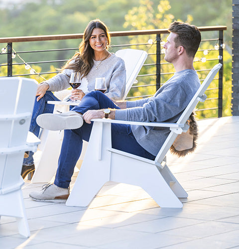 A man and woman sit on a sunlit patio in white Adirondack chairs, holding glasses of red wine. They are smiling and engaged in conversation. The background features a railing with string lights and a view of greenery.