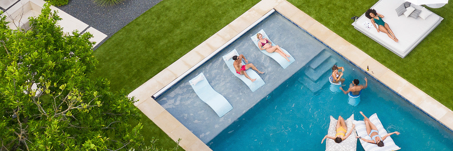 Aerial view of a modern backyard with a rectangular pool. People are lounging on poolside chairs and in the water on floaties. Nearby, two people relax on a sunbed. Green grass and a tree border the pool area.
