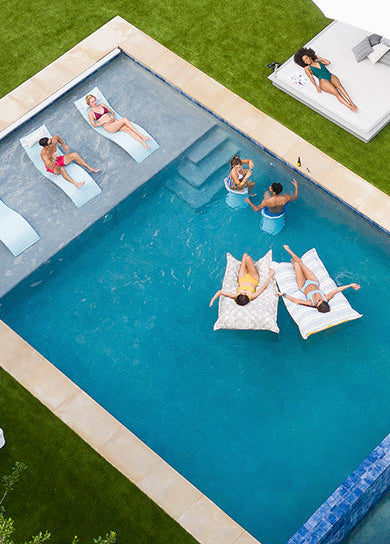 Aerial view of six people enjoying a pool on a grassy lawn. Four people are in the pool on lounge chairs and inflatables, while two are sunbathing on a nearby raised platform under a white umbrella.