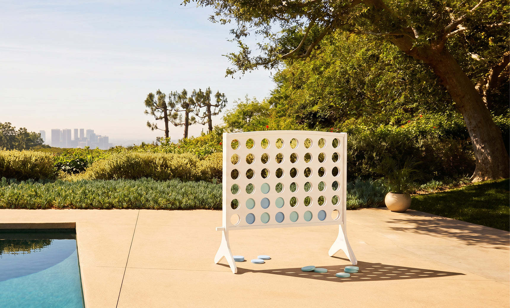 The Ledge Lounger Four In A Row stands on a patio by the pool, surrounded by lush greenery and trees, with a distant city skyline under a clear blue sky.
