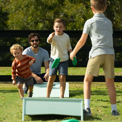 A man sits on the grass, while three boys play cornhole. One boy tosses a green bean bag towards a wooden board. The group is outdoors, with a black fence and trees in the background.