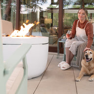A person relaxes on a patio chair, holding a glass of wine and petting a dog. The Ledge Lounger Halo Firepit, with its pivot-glide lid, emits a warm glow, enhancing the cozy outdoor ambiance on the wooden deck.