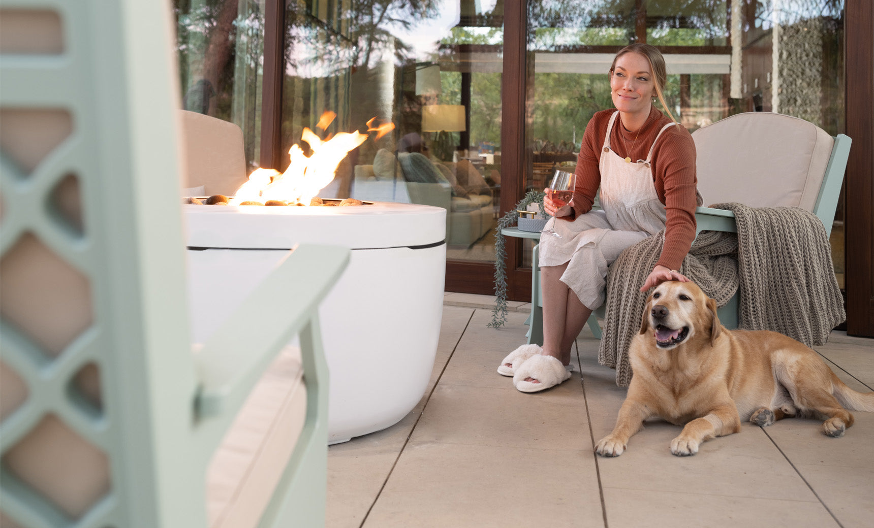 A person relaxes on a patio chair, holding a glass of wine and petting a dog. The Ledge Lounger Halo Firepit, with its pivot-glide lid, emits a warm glow, enhancing the cozy outdoor ambiance on the wooden deck.