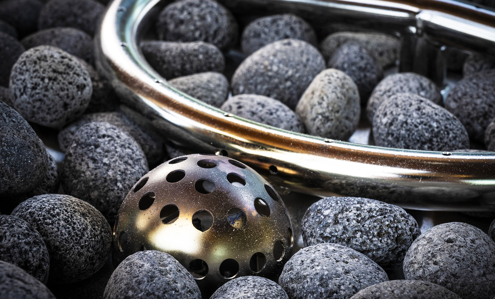 A collection of smooth gray stones encircles a circular metallic object and a dome-like perforated piece, creating an elegant propane fire pit setup. This outdoor ambiance is reminiscent of the Halo Firepit by Ledge Lounger.