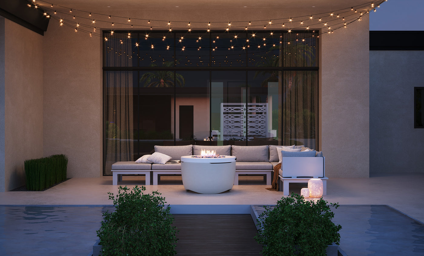 A modern patio with a Ledge Lounger Halo Firepit surrounded by a sectional sofa and illuminated by string lights. A small pool, bordered by potted plants, sits in the foreground, while large windows with sheer curtains cast gentle light on this serene setting.