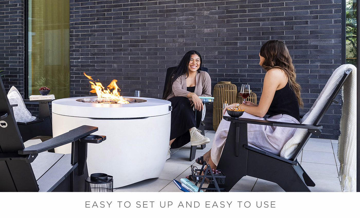 Two people sit on patio chairs with Ledge Loungers round Halo Firepit, enhancing the outdoor ambiance. One holds a glass of wine as they smile and chat against a dark brick wall. Caption: EASY TO SET UP AND EASY TO USE.