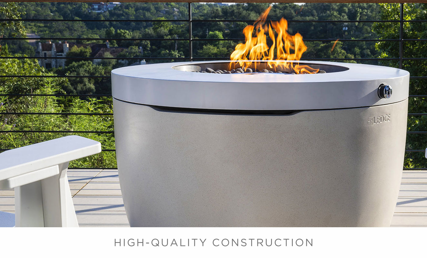 A modern outdoor patio features the sleek Halo Firepit by Ledge Lounger, with a pivot-glide lid and surrounded by a white chair. This serene setup enhances the ambiance, overlooking greenery framed by trees, with HIGH-QUALITY CONSTRUCTION prominently displayed.