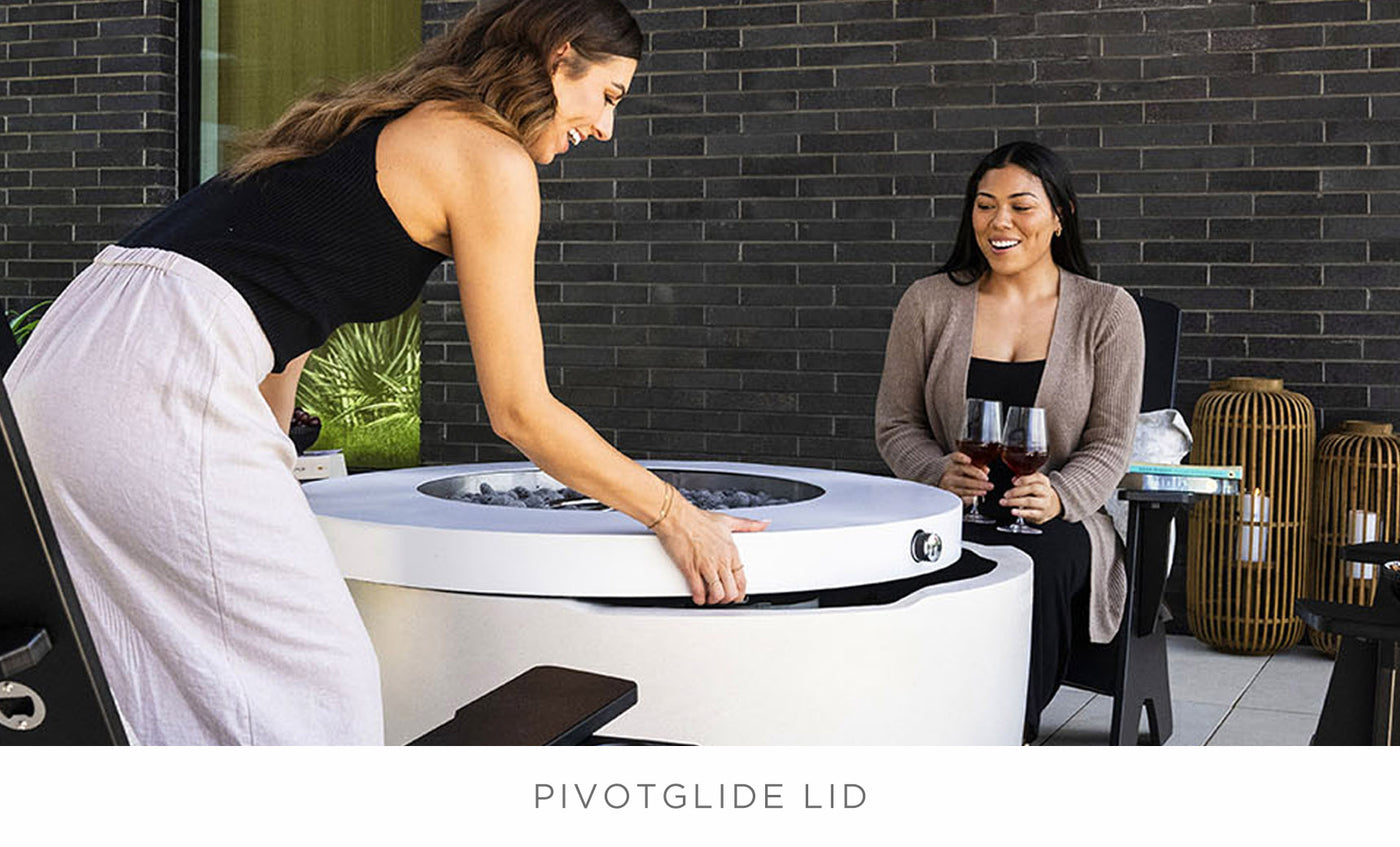 Two women enjoy the outdoors with a Halo Aflame Bundle by Ledge Lounger. One adjusts its lid, while the other sips wine. Theyre surrounded by modern patio furniture and decorative lanterns.