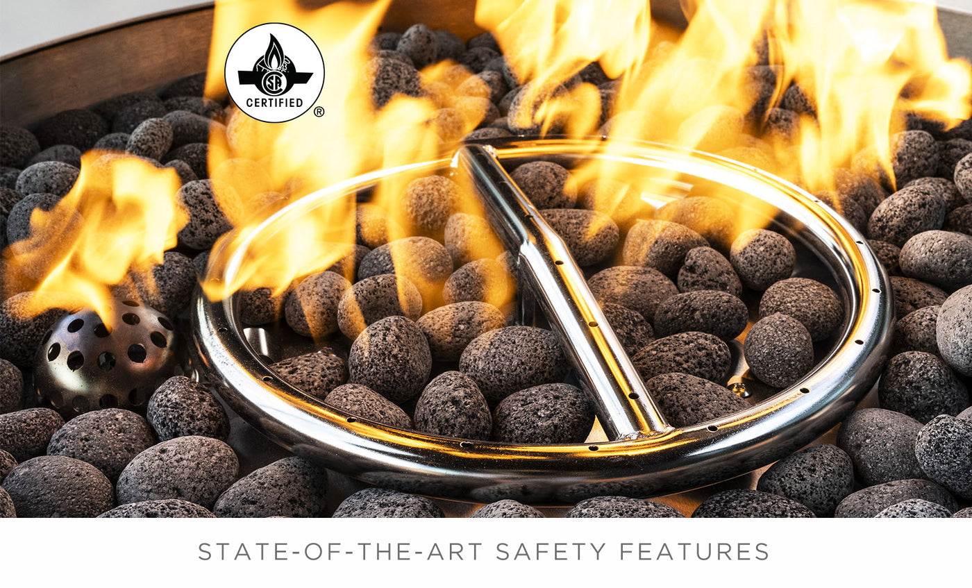 The Halo Aglow Bundle by Ledge Lounger features a circular gas fire pit with flames dancing over black lava rocks and a stainless steel burner ring. In the top left corner, it displays a certification logo. The bottom text highlights State-of-the-art safety features.