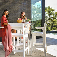 Two people sit on Ledge Loungers Haven Hightop Set on a terrace by a pool with a glass railing. One wears red, the other a patterned dress. A bowl of fruit sits on the table as trees and blue sky form the backdrop.