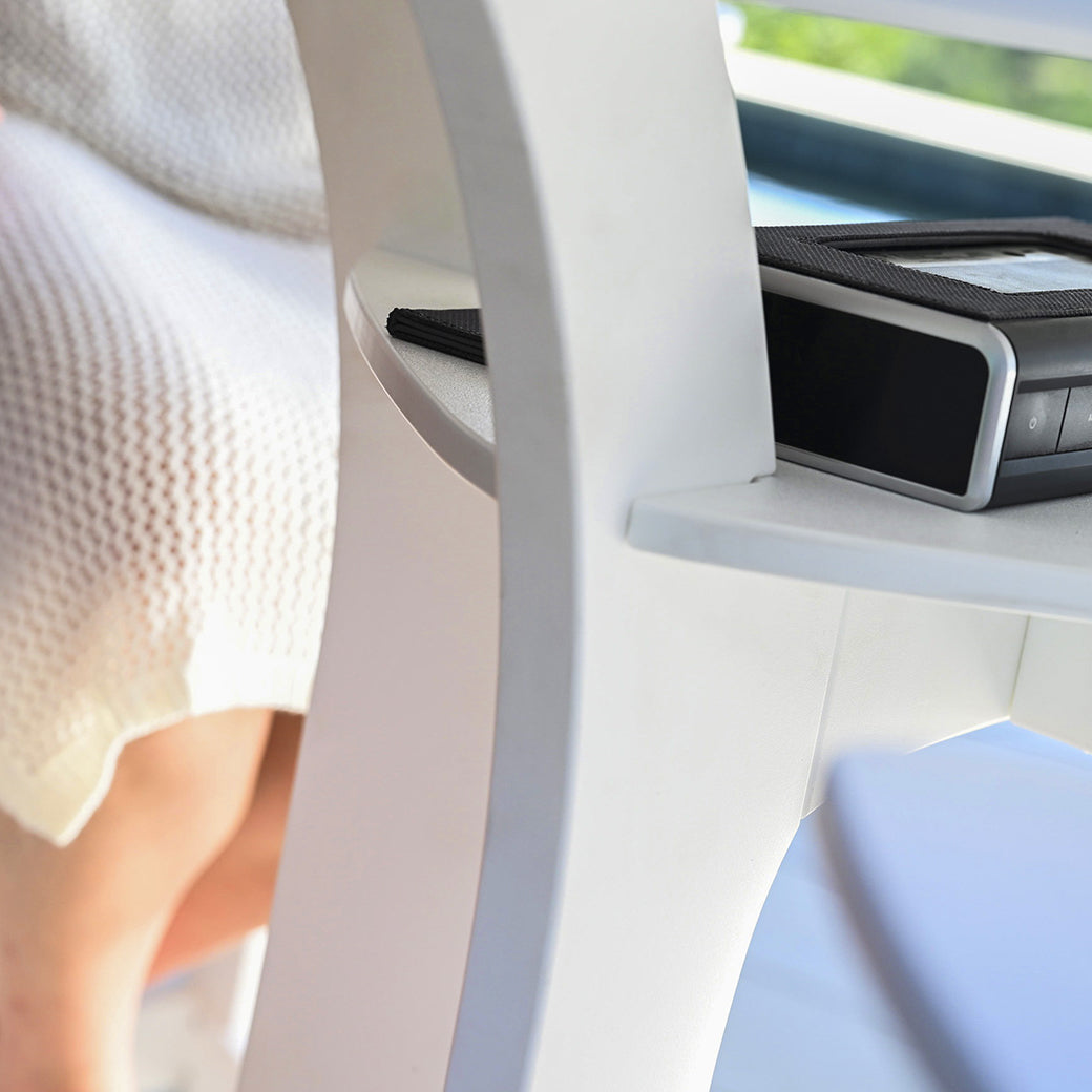 Two people relax by the brightly lit Haven Hightop Table from Ledge Lounger. One wears a light dress, the other has bare legs, while a black electronic device with buttons is on the table.