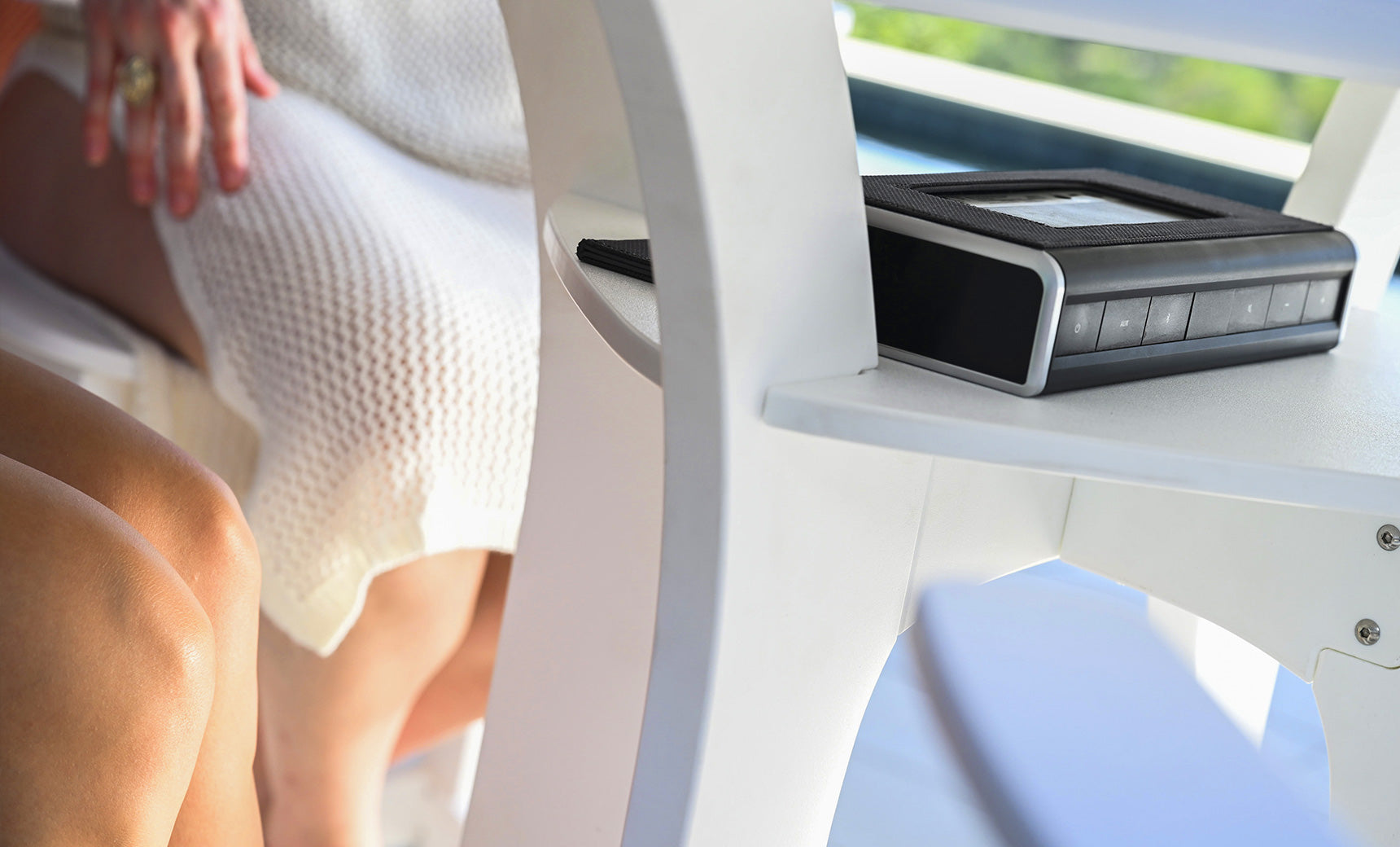 Two people relax by the brightly lit Haven Hightop Table from Ledge Lounger. One wears a light dress, the other has bare legs, while a black electronic device with buttons is on the table.