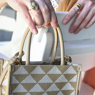 A person holds a patterned handbag with beige and white geometric triangles, wearing a ring and a gold bracelet. In the background, theres a white Haven Hightop Set by Ledge Lounger and soft pink fabric draped beside them.