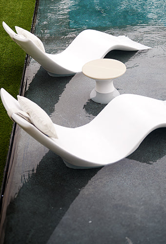 Two modern, white, wavy lounge chairs with cushions are partially submerged in a pool next to a small round table. Green grass borders the pool area, creating a tranquil outdoor setting.