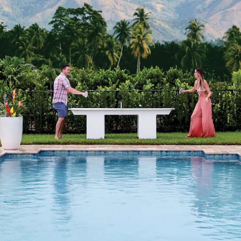 A man and woman are playing table tennis outdoors beside a swimming pool. The background features lush greenery with palm trees and mountains. The woman is wearing a long pink dress, and the man is in a plaid shirt and shorts.
