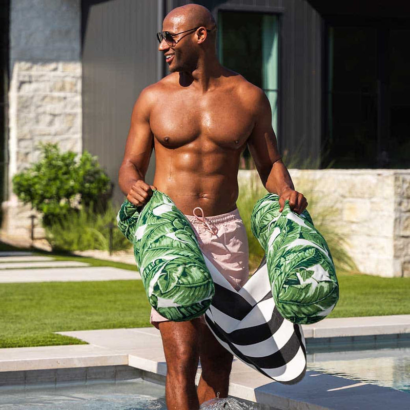 A smiling man in pink swim trunks stands by a pool holding two large tropical leaf-patterned pool floats, with a house and greenery in the background.