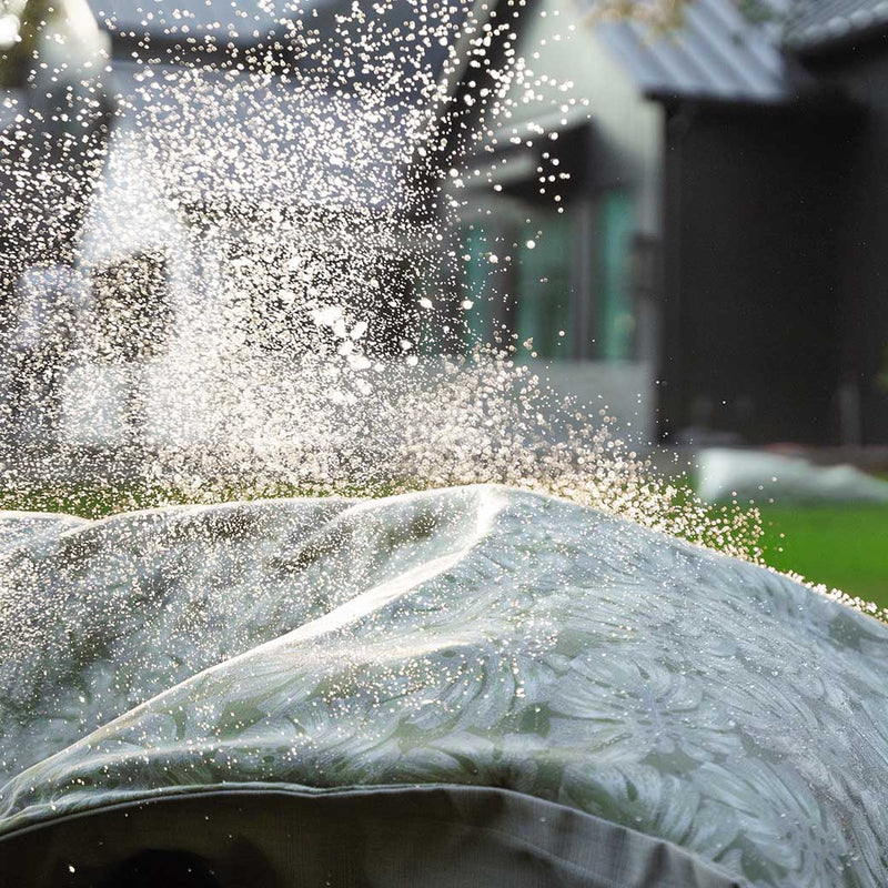 Water droplets splash in the air, captured in motion over a green and gray patterned outdoor cushion. The background is blurred, showing a hint of buildings and greenery on a sunny day.