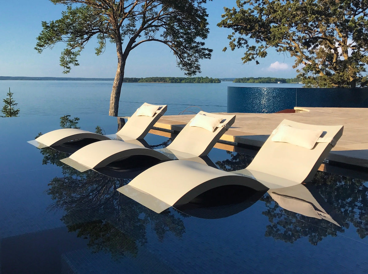 Beige lounge chairs on a wooden deck by a body of water with trees in the background.
