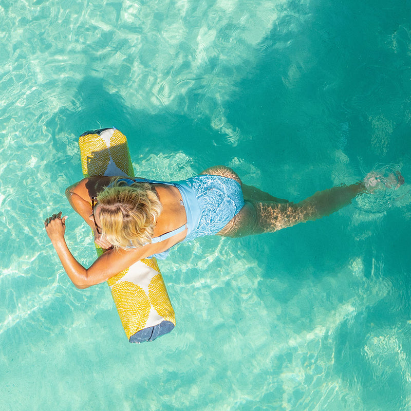A person with blonde hair wearing a blue swimsuit floats on a colorful inflatable raft in a clear turquoise pool. The sunlight creates shimmering patterns on the water.