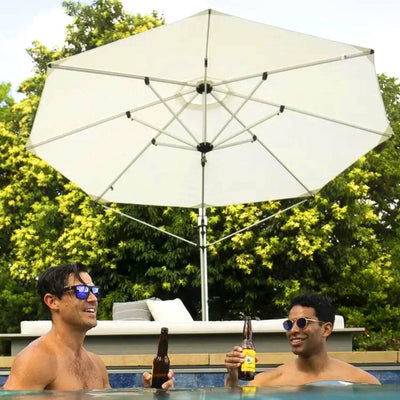 Two people are enjoying drinks in a pool under a large umbrella on a sunny day. They are wearing sunglasses and smiling at each other. Green trees are visible in the background, creating a relaxed, summer atmosphere.