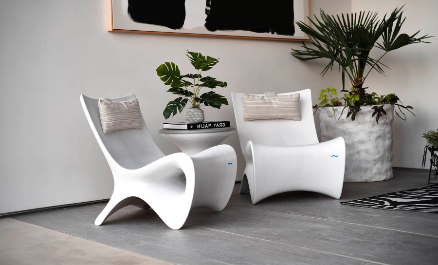 Two modern white chairs with pillows are positioned in a stylish room. A small table between them holds a book and a potted plant. A large planter with green plants is in the background, and a monochrome abstract artwork hangs on the wall.