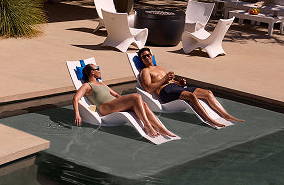 Two people reclining on white outdoor lounge chairs beside a swimming pool on a patio.